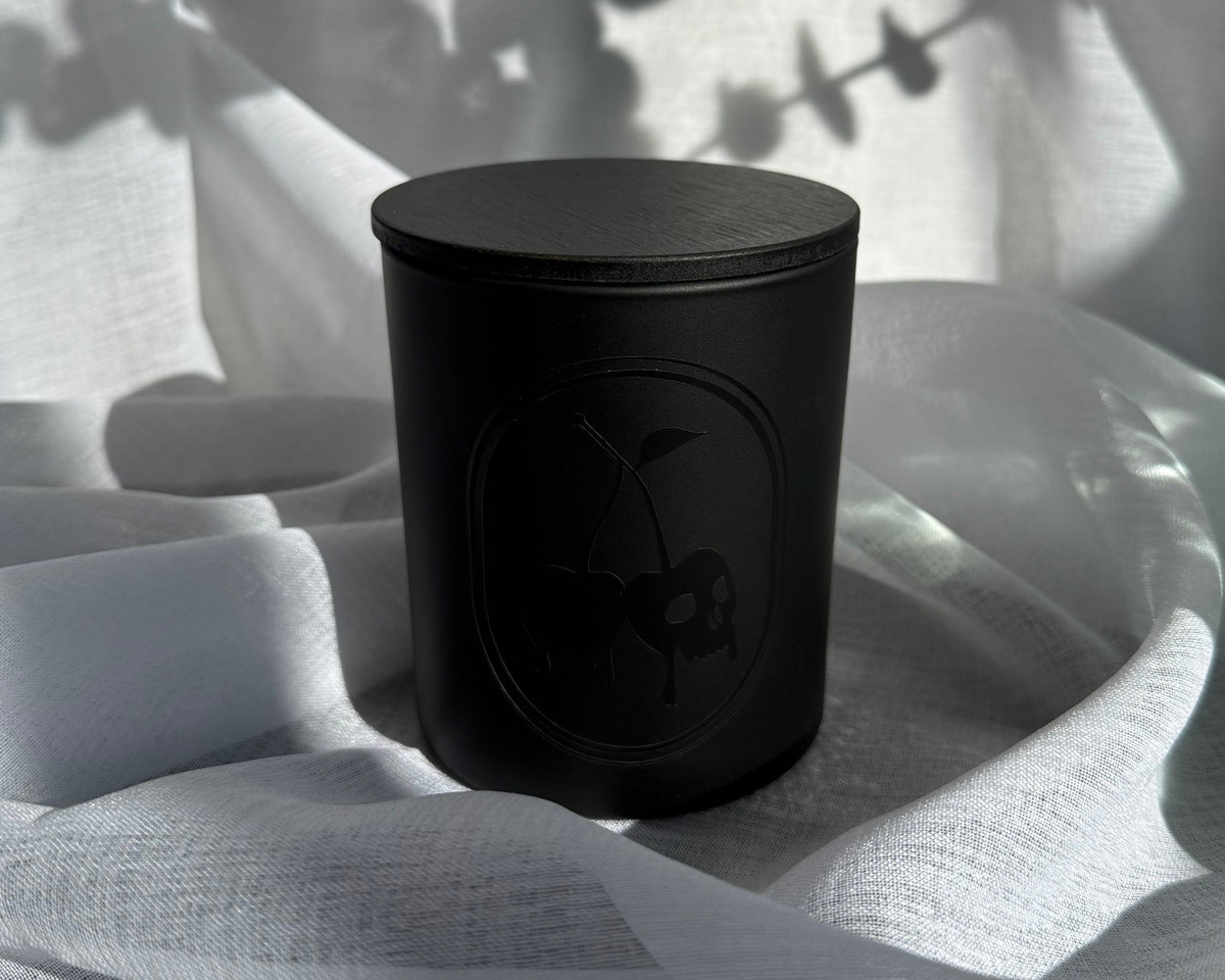 A matte black cylindrical candle jar with a matching lid, featuring an embossed floral emblem on the front, set against draped white fabric with soft leaf shadows.
