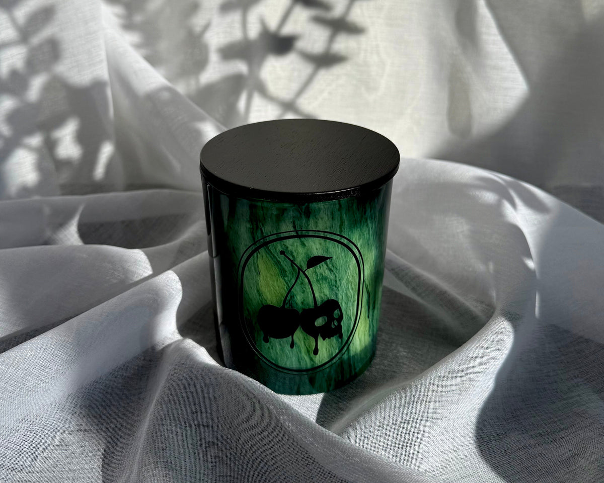 A small cylindrical candle jar with a black lid and a green marbled body featuring a black ant illustration, set on draped white fabric with leaf shadows cast across it.