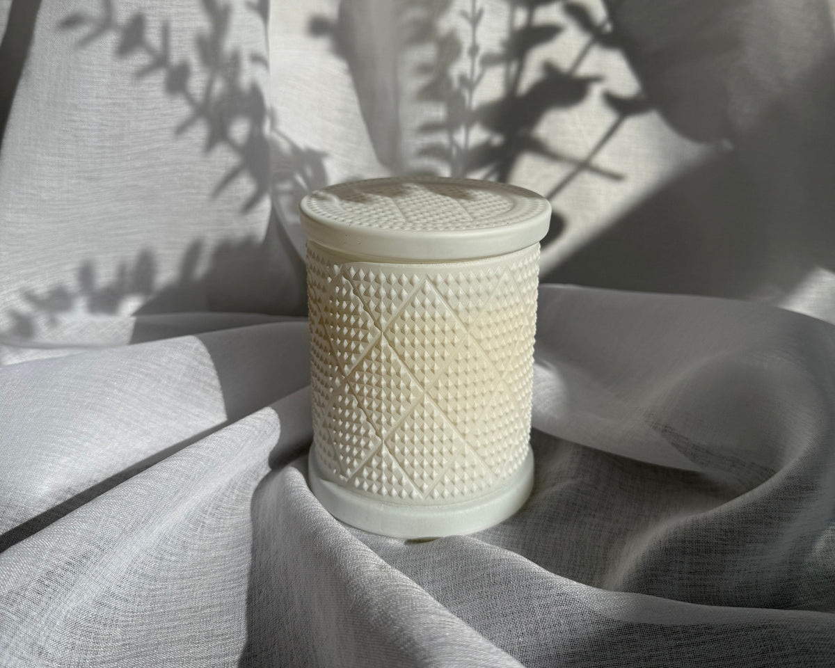 A textured candle in an white ceramic container with a matching lid, set against draped white fabric with leaf shadows cast across it.