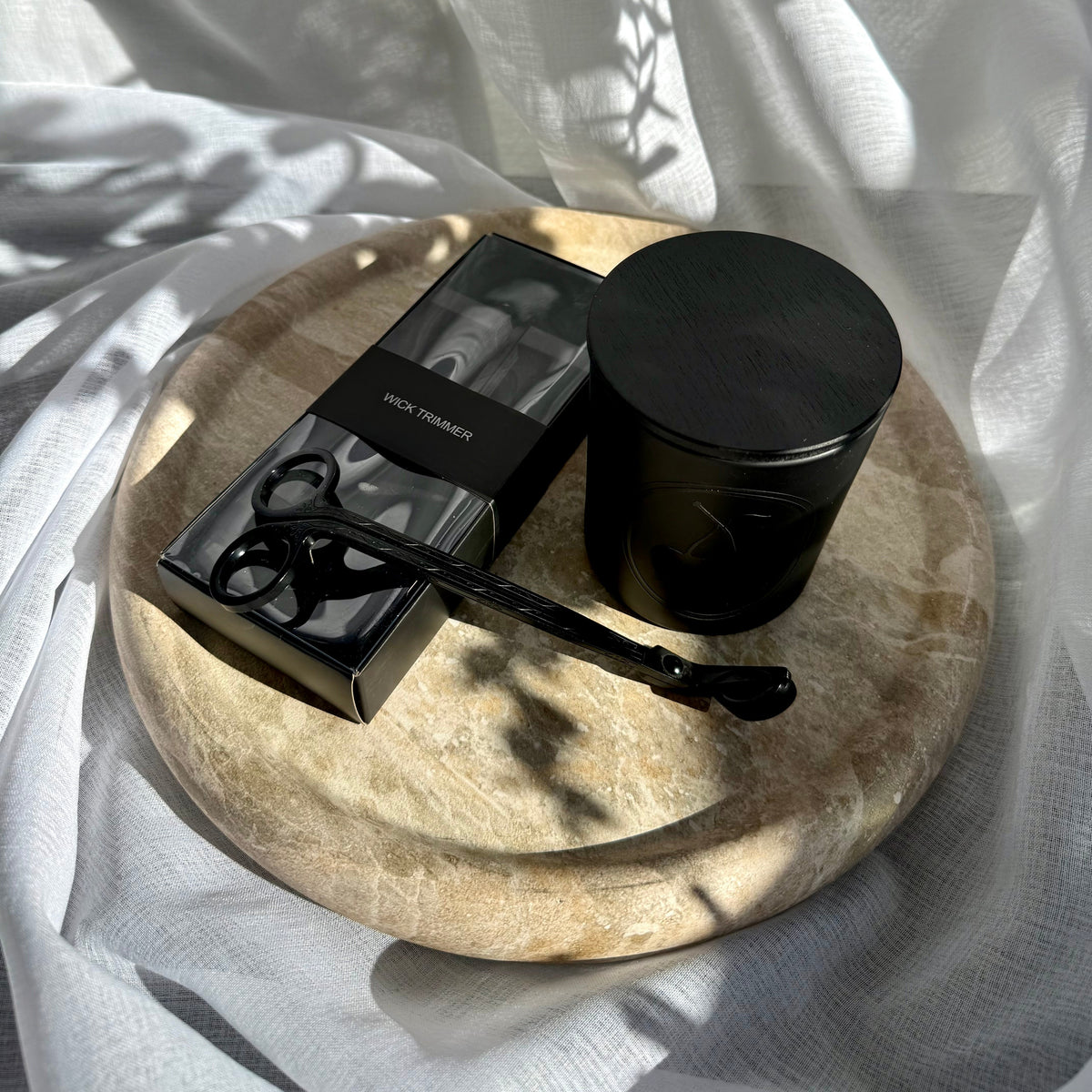 A black candle and a black rectangular box with a black wick trimmer, arranged on a round stone tray draped with white fabric under dappled sunlight.