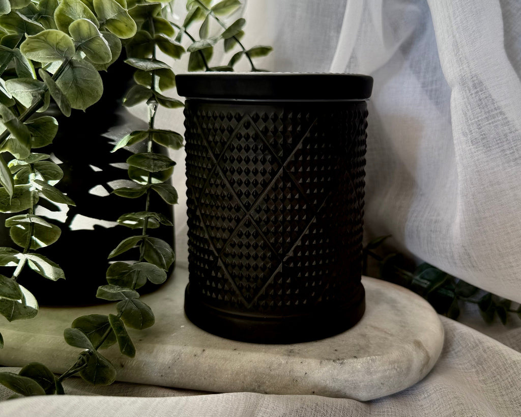 A black textured candle holder with a diamond pattern, styled on a marble coaster with a green plant and white fabric drape in the background.