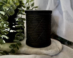 A black textured candle holder with a diamond pattern, styled on a marble coaster with a green plant and white fabric drape in the background.