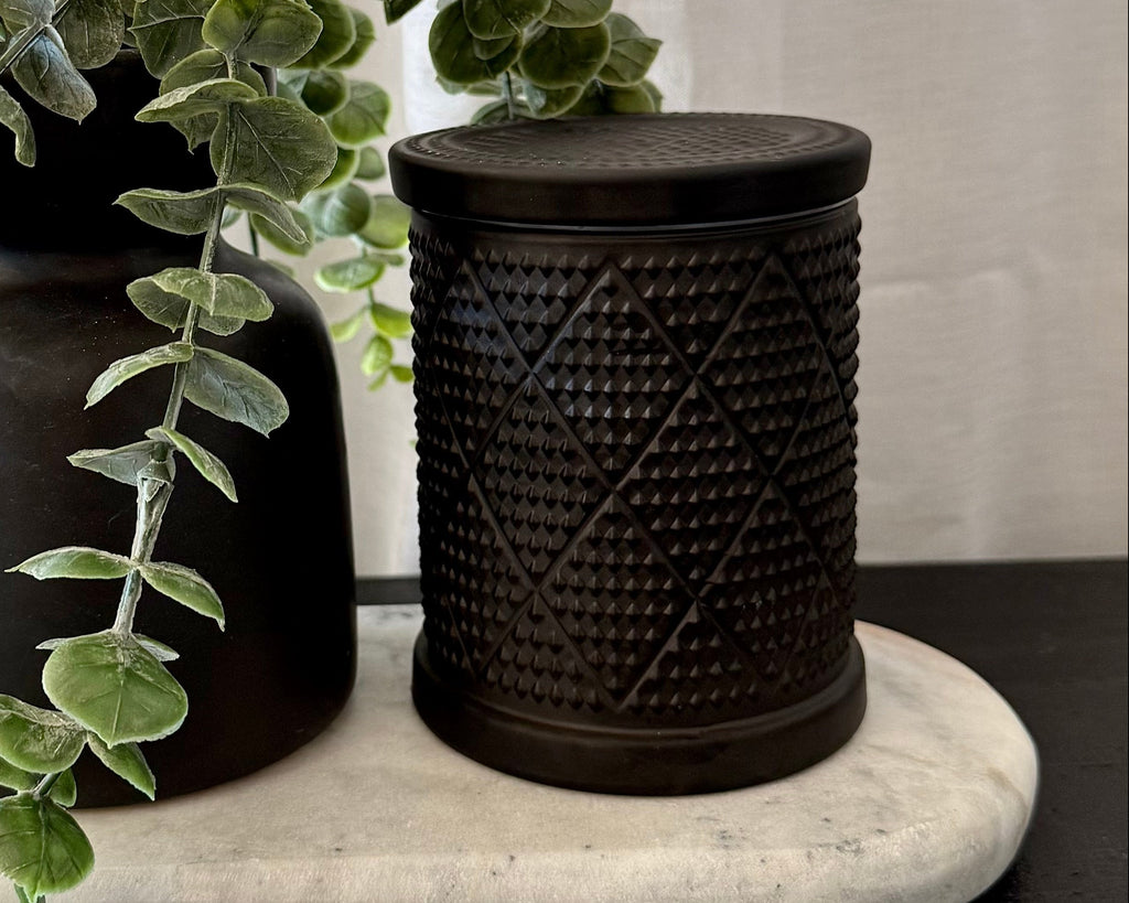 One matte black vase with trailing green eucalyptus and one textured black cylindrical jar with a lid, displayed on a white marble tray over a dark wooden surface with a white curtain background.