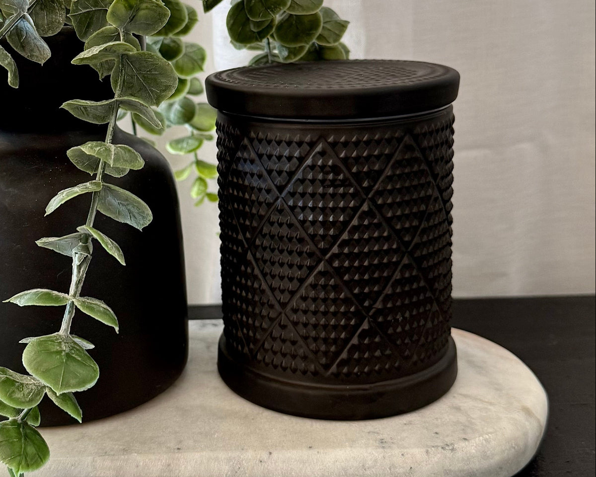 One matte black vase with trailing green eucalyptus and one textured black cylindrical jar with a lid, displayed on a white marble tray over a dark wooden surface with a white curtain background.