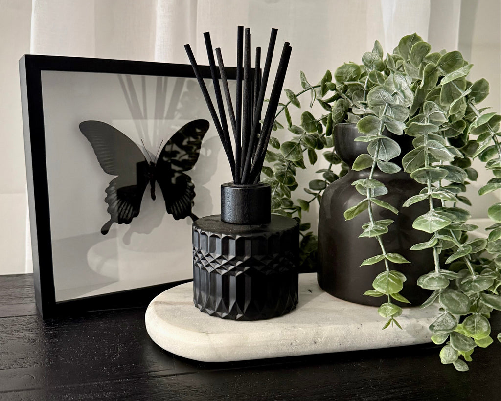 A black reed diffuser with black reeds sits on a white marble tray next to a black ceramic vase with a green trailing plant, with a black-framed butterfly print in the background.
