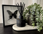 A black reed diffuser with black reeds sits on a white marble tray next to a black ceramic vase with a green trailing plant, with a black-framed butterfly print in the background.