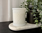 A white textured ceramic candle with a matching lid, displayed on a white marble tray next to a matte black vase with green eucalyptus leaves, set on a dark wooden table against a white curtain background.