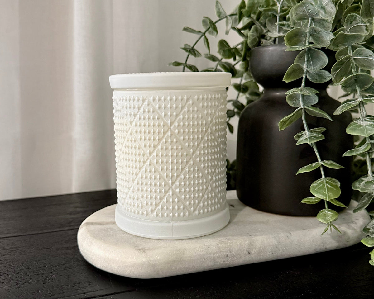 A white textured ceramic candle with a matching lid, displayed on a white marble tray next to a matte black vase with green eucalyptus leaves, set on a dark wooden table against a white curtain background.