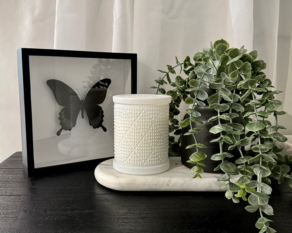 A white textured ceramic candle jar sits on a white marble tray next to a black-framed butterfly print and a green trailing eucalyptus plant on a dark wooden table, with white sheer curtains in the background.
