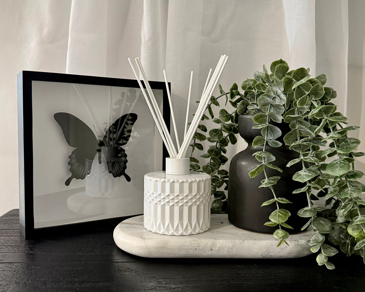 A styled interior scene showing a white textured reed diffuser with white reeds on a marble tray, next to a dark ceramic vase with green eucalyptus leaves and a framed butterfly print, all set on a dark wooden surface with white sheer curtains in the background.