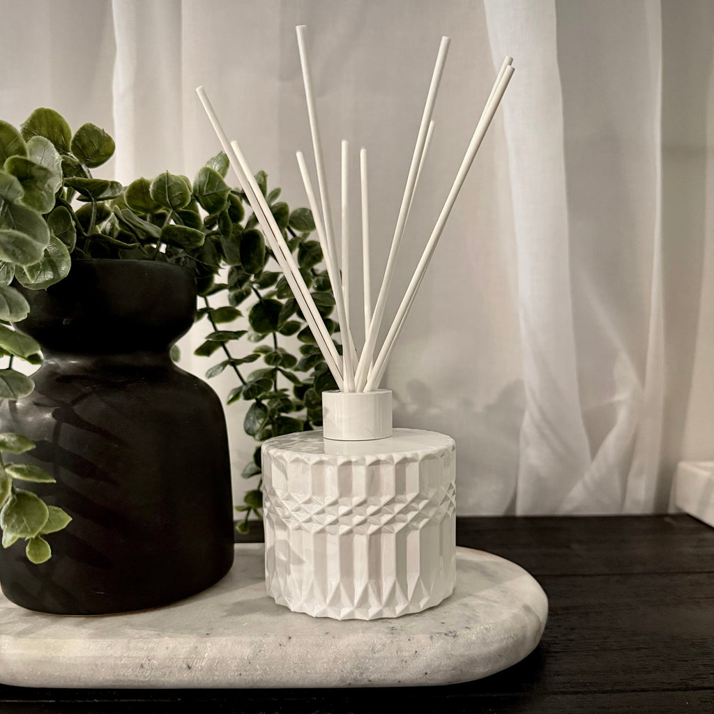 A white textured ceramic reed diffuser with white reeds on a marble tray, styled next to a black vase with green foliage against a white curtain background.