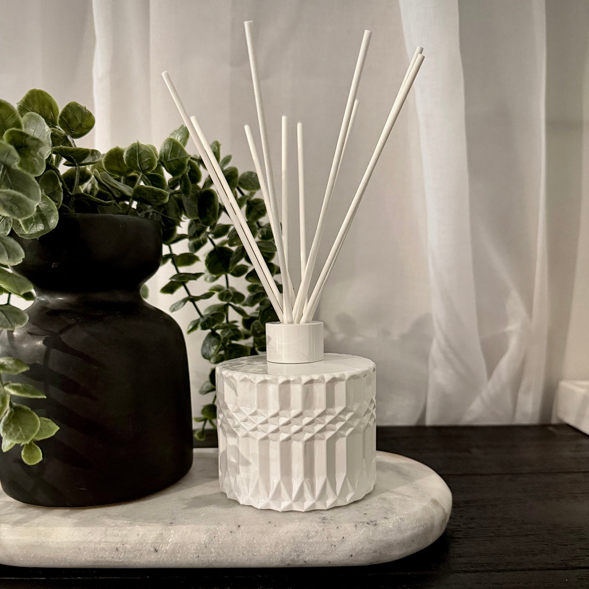A white textured ceramic reed diffuser with white reeds on a marble tray, styled next to a black vase with green foliage against a white curtain background.