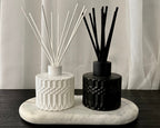 Two reed diffusers, one white and one black, displayed on a white marble tray on a dark wooden table in front of white sheer curtains.