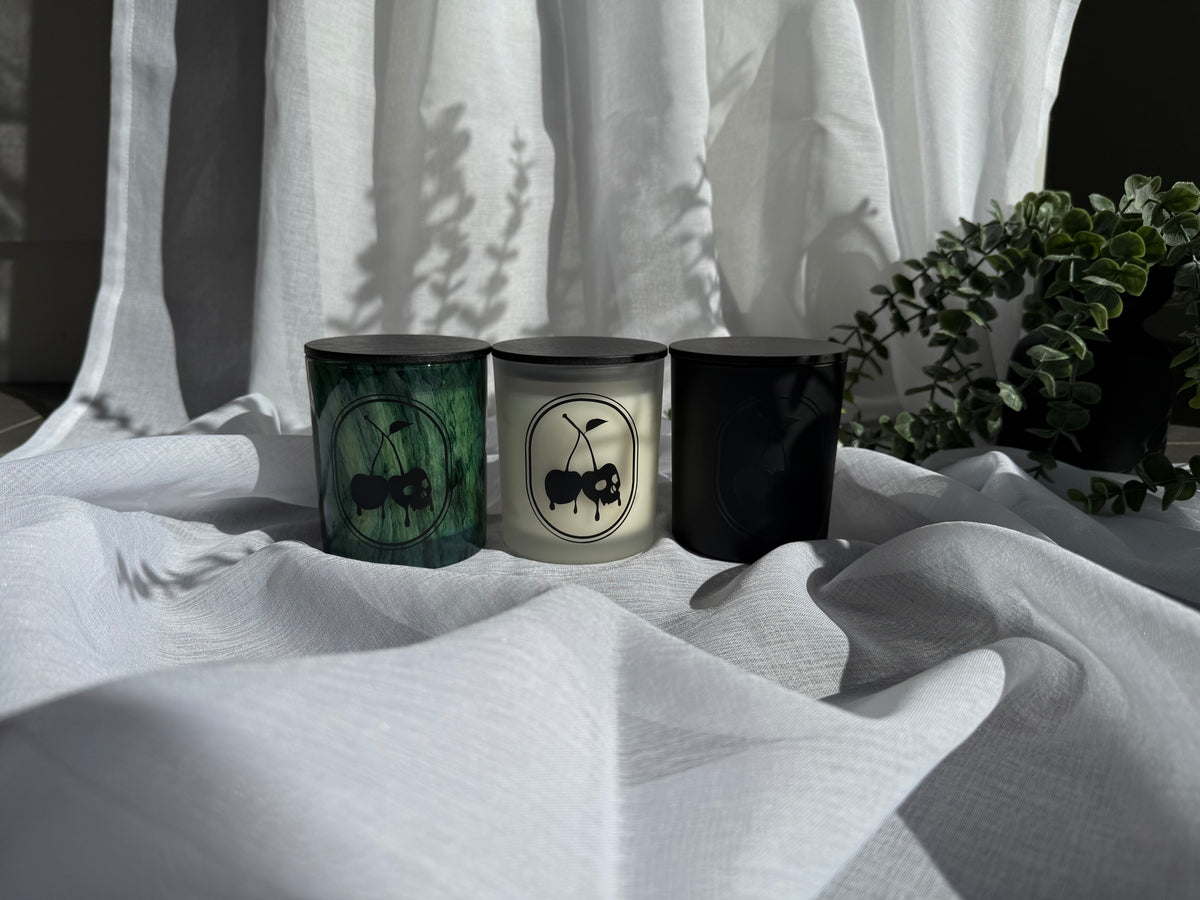 Three decorative candles in cylindrical glass jars with black lids are arranged on draped white fabric. The left candle has a dark green marbled pattern with a black ant illustration, the center candle is white with a black cherry illustration, and the right candle is solid black.