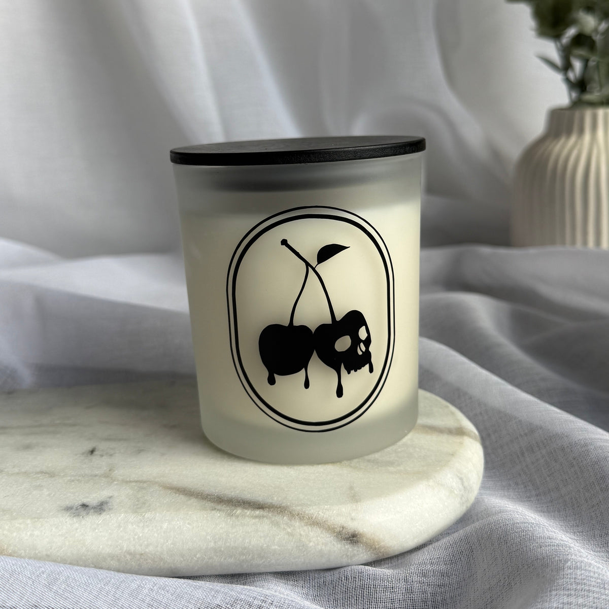 A frosted glass candle jar with a black lid and a black cherry logo on the front, set on a marble coaster against draped white fabric with a blurred vase in the background.