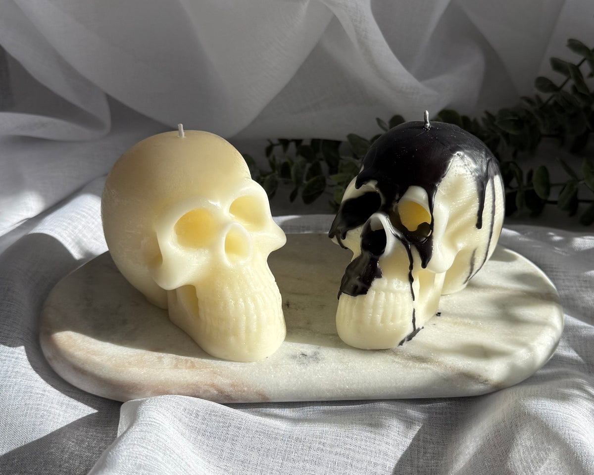 Two decorative candles displayed on a small stone tray: one sculpted skull candle in warm cream and one animal-shaped candle in black, cream, and white with yellow drips. The candles are styled on draped white fabric with soft shadows and a green plant in the background.
