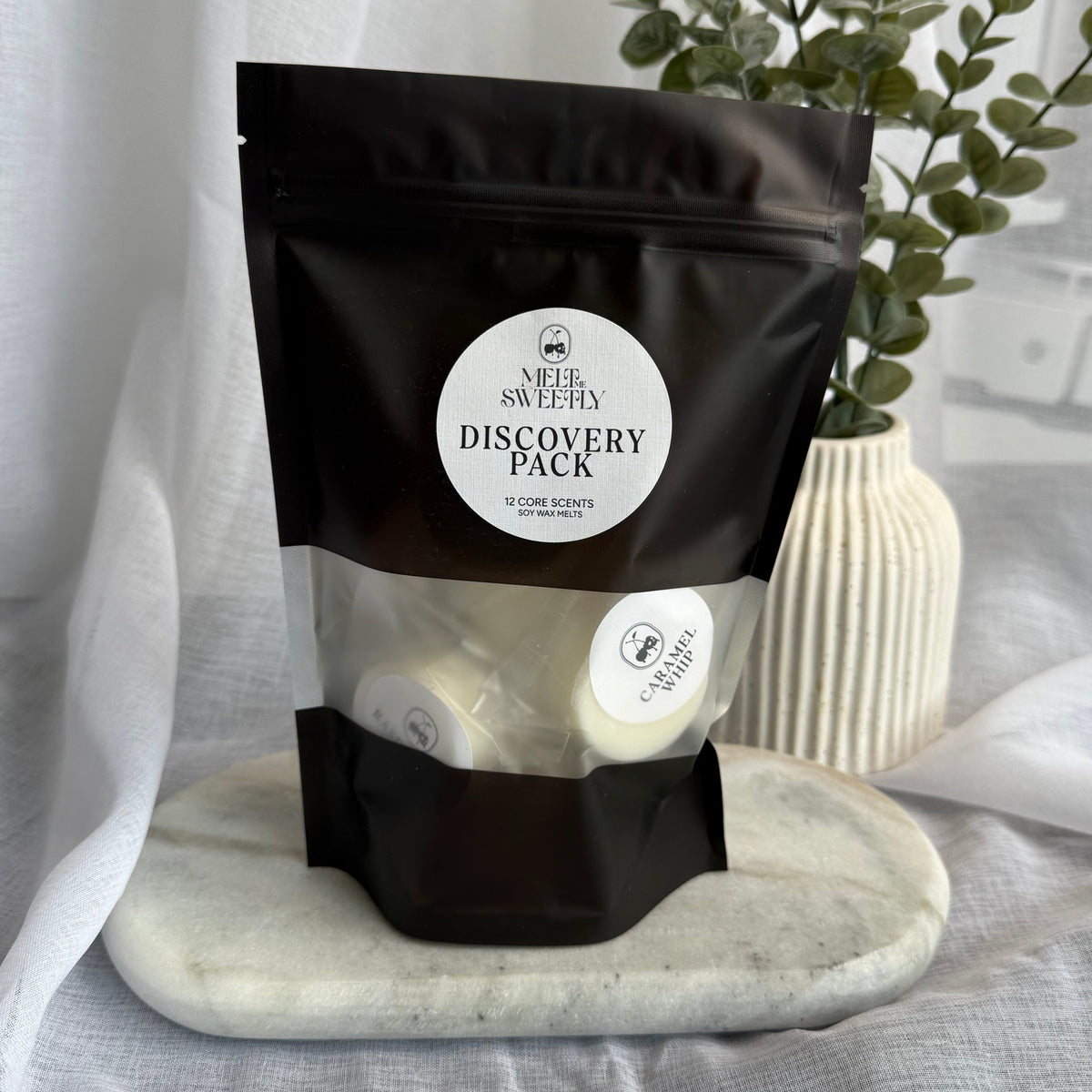 A black resealable pouch labeled 'DISCOVERY PACK' with a clear window showing white wax melts inside, styled on a marble tray with draped white fabric and a potted green plant in the background.