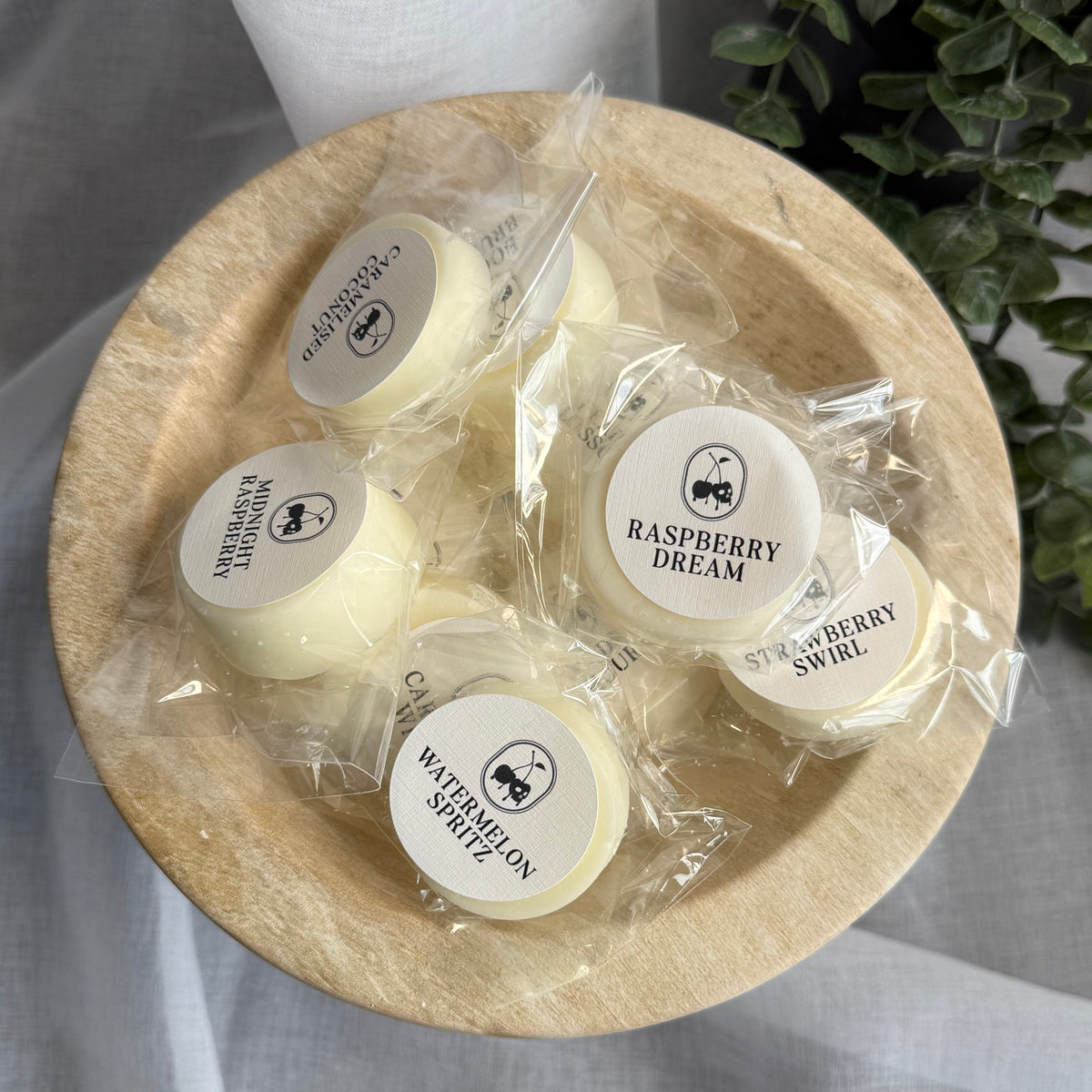 Small wrapped cream-colored wax melts or soaps with round white labels are arranged in a shallow round wooden bowl on draped light gray fabric, with a green leafy plant in the background.