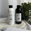 Two bottles of diffuser refill, one in white liquid gold and one in black strawberry swirl, displayed on a marble slab with a green plant in the background.