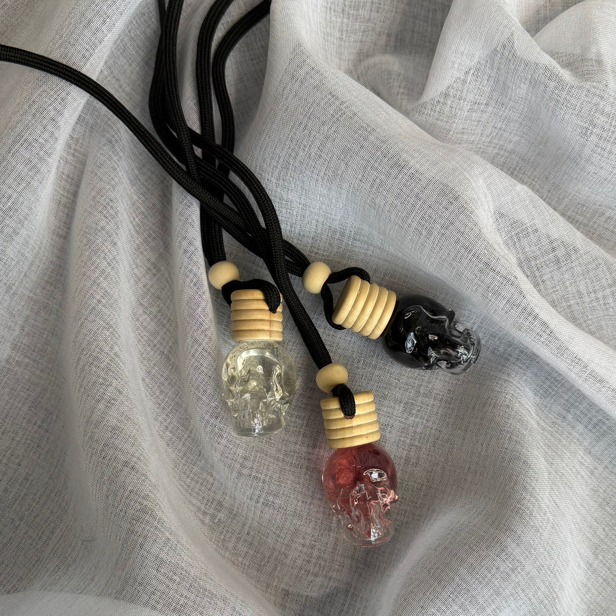 Three crystal skull pendant necklaces with black cords and beige spacer beads, displayed on a textured white fabric background.