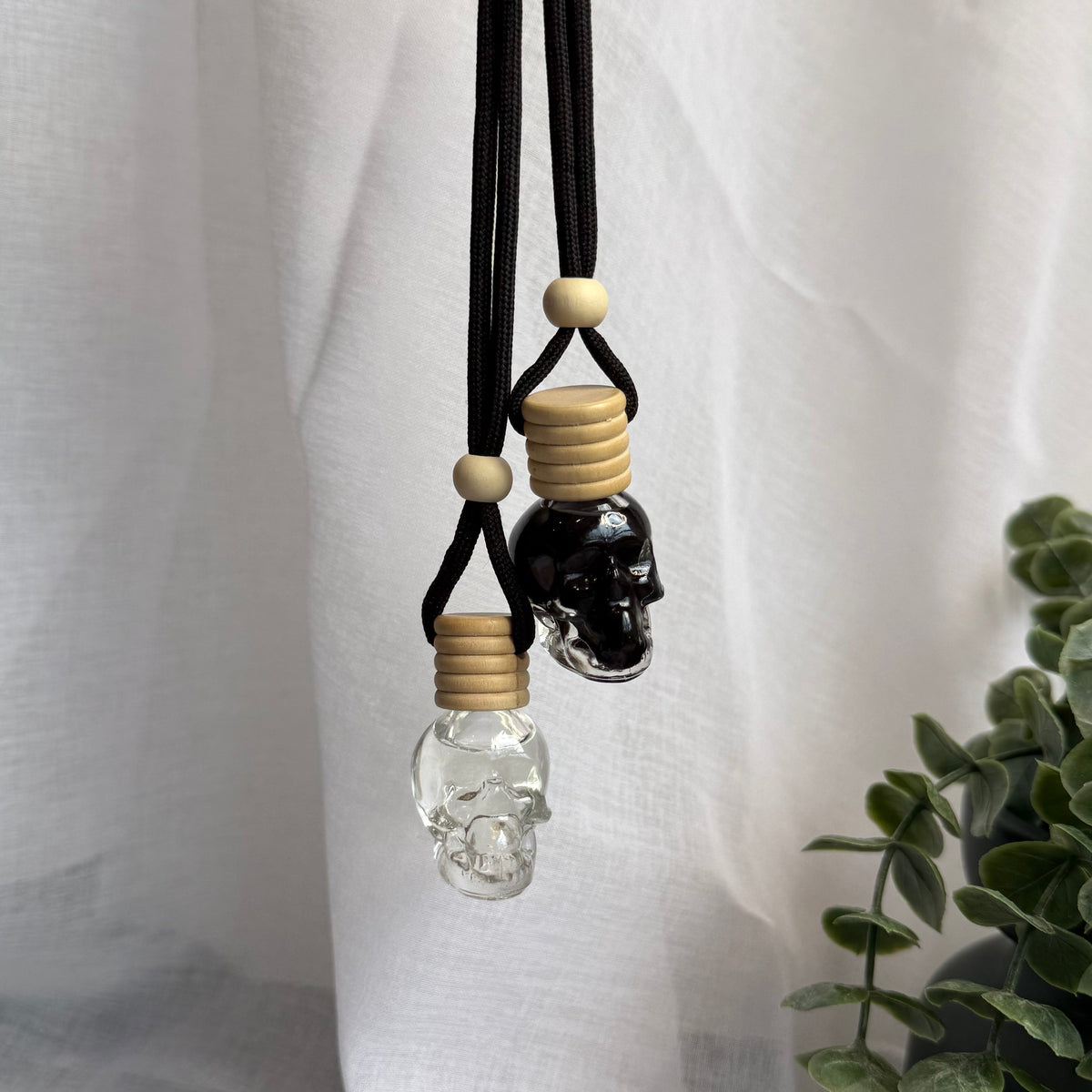 Two skull pendant necklaces on black cords, one with a clear glass skull and one with a black skull, both featuring light wood caps and beads, held by a hand against a white draped fabric backdrop with a green plant in the corner.