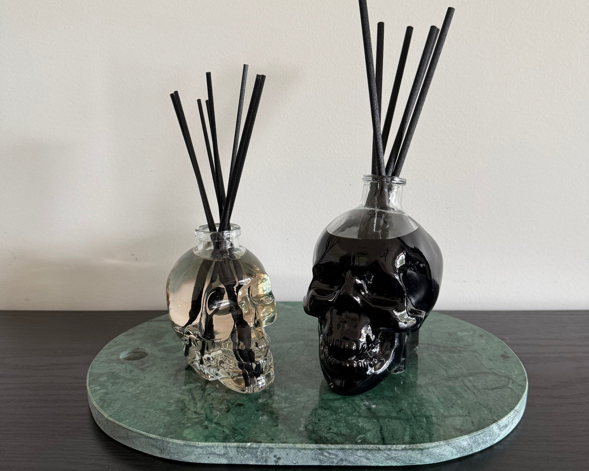 Two skull-shaped reed diffusers on a round green marble tray, set on a dark wooden surface against a plain off-white wall.