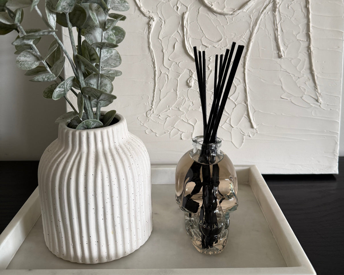 A styled interior scene showing a white ribbed ceramic vase with eucalyptus branches and a clear glass reed diffuser bottle with black reeds, both placed on a white tray against a textured white wall.
