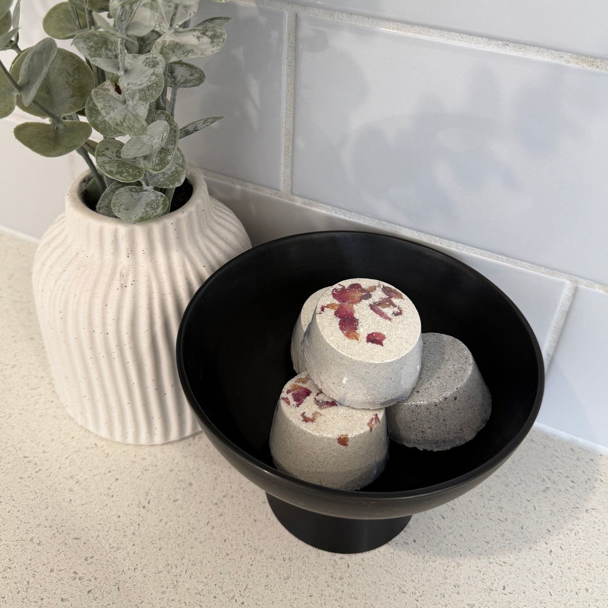 Three cylindrical Shower steamers with white and pink botanical patterns are arranged inside a black bowl, which is placed on top of a ribbed white ceramic vase. The setup is styled on a speckled beige countertop next to a white tiled wall with a green eucalyptus branch in the foreground.